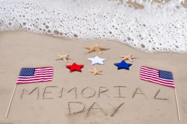 "Memorial Day carved out on beach sand with flags and blue, white, and red stars"